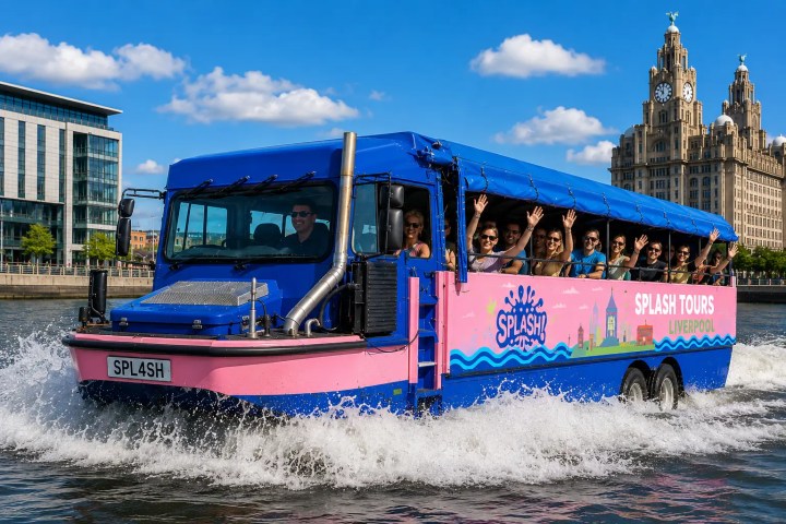Tour group on pink and blue amphibious bus on water with waving passengers, Liverpool landmarks in the background.