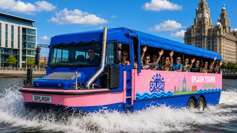 Tour group on pink and blue amphibious bus on water with waving passengers, Liverpool landmarks in the background.