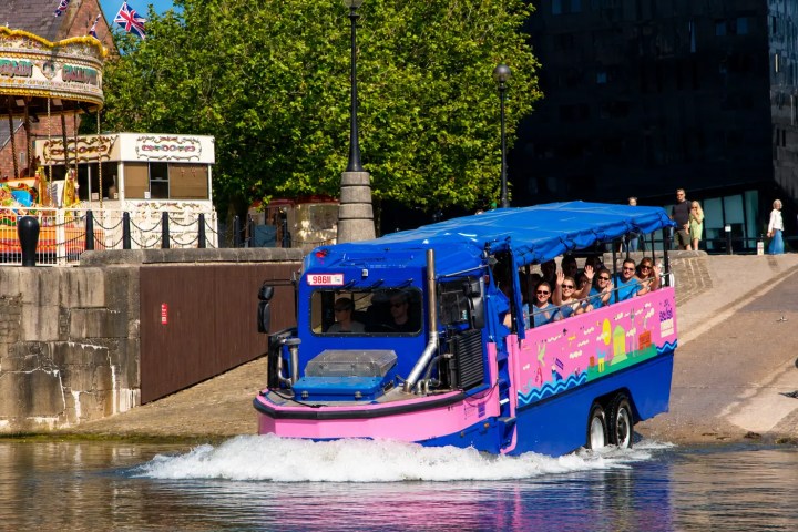 Amphibious tour bus entering water near merry-go-round and trees.