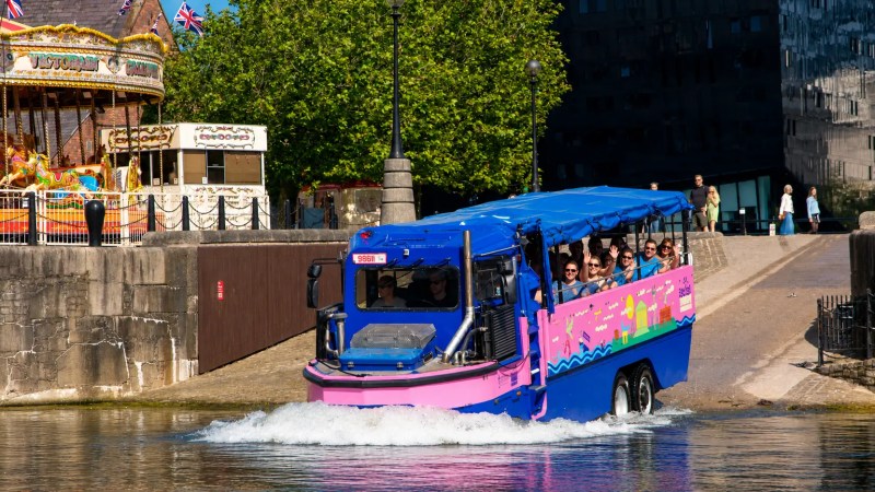 Amphibious tour bus entering water near merry-go-round and trees.